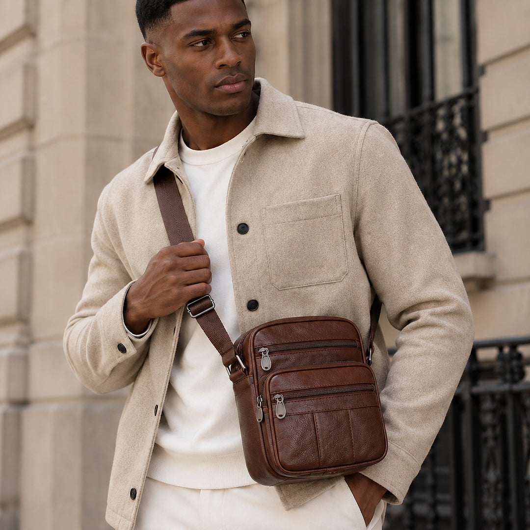 A man wearing a beige overshirt and white t-shirt carries a brown leather crossbody bag with a matching brown strap, standing in front of a stone building.