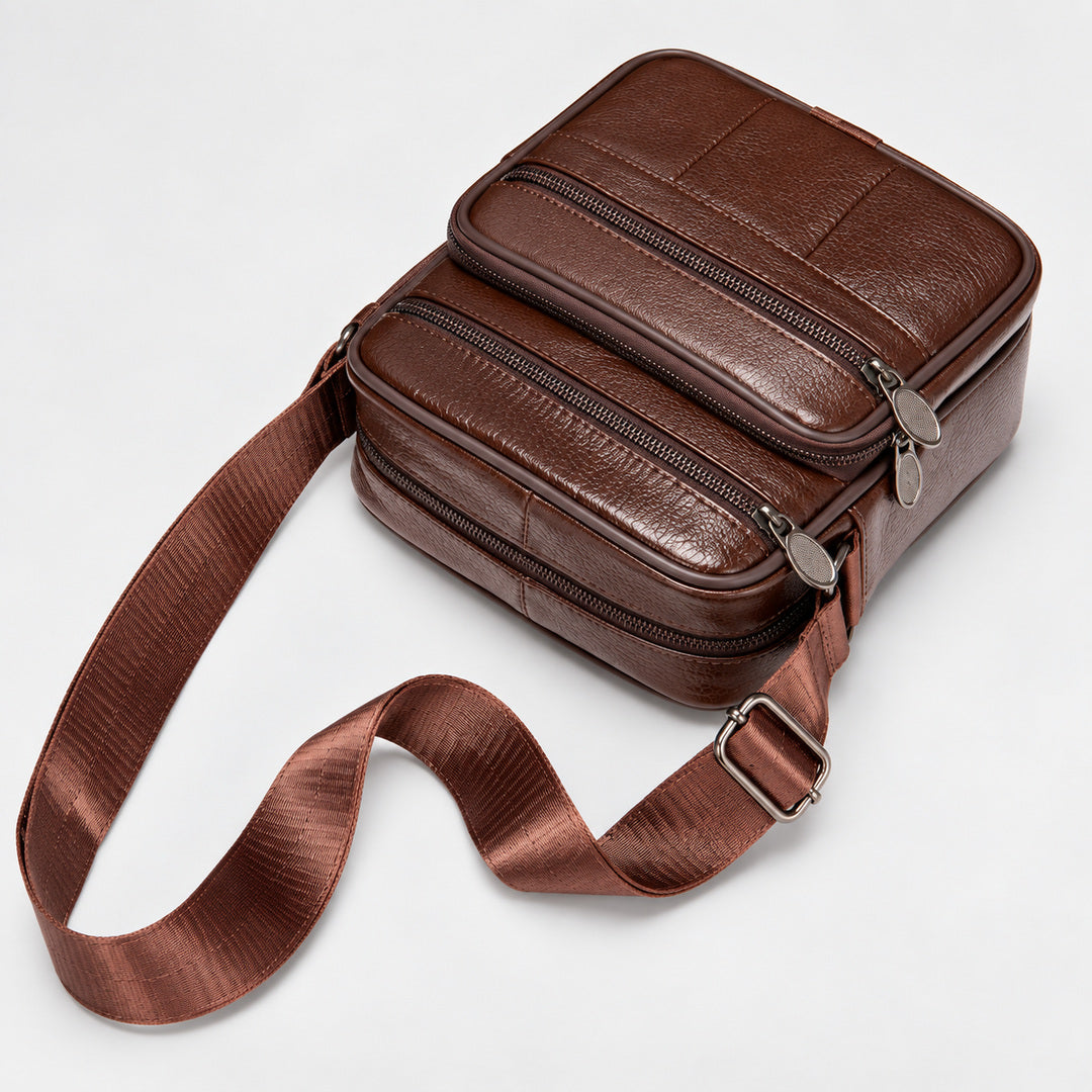 A brown leather crossbody bag with multiple zippered compartments and a wide matching leather strap, photographed on a clean light gray background.