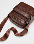 A brown leather crossbody bag with multiple zippered compartments and a wide matching leather strap, photographed on a clean light gray background.
