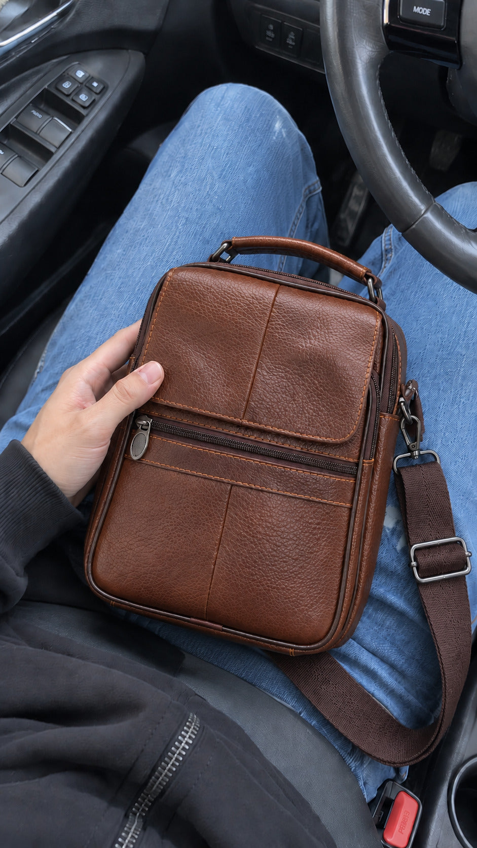 A brown leather crossbody bag with a top handle and adjustable shoulder strap, held by a person sitting in a car. The person is wearing blue jeans and a dark gray hoodie.