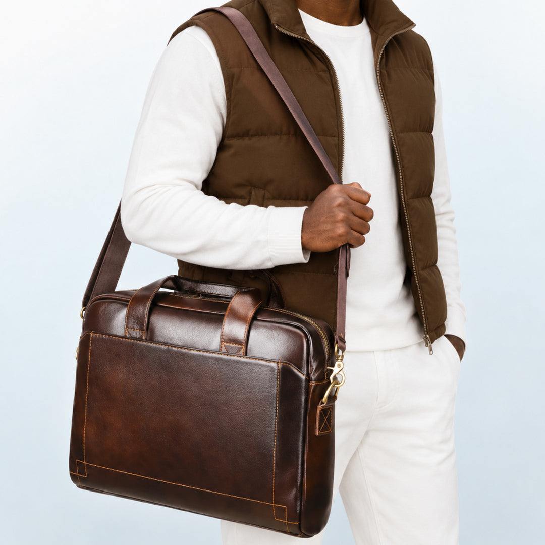 A man wearing a brown puffer vest, white long-sleeve shirt, and white pants carries a dark brown leather briefcase-style shoulder bag with a matching strap against a light blue studio background.