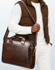 A man wearing a brown puffer vest, white long-sleeve shirt, and white pants carries a dark brown leather briefcase-style shoulder bag with a matching strap against a light blue studio background.