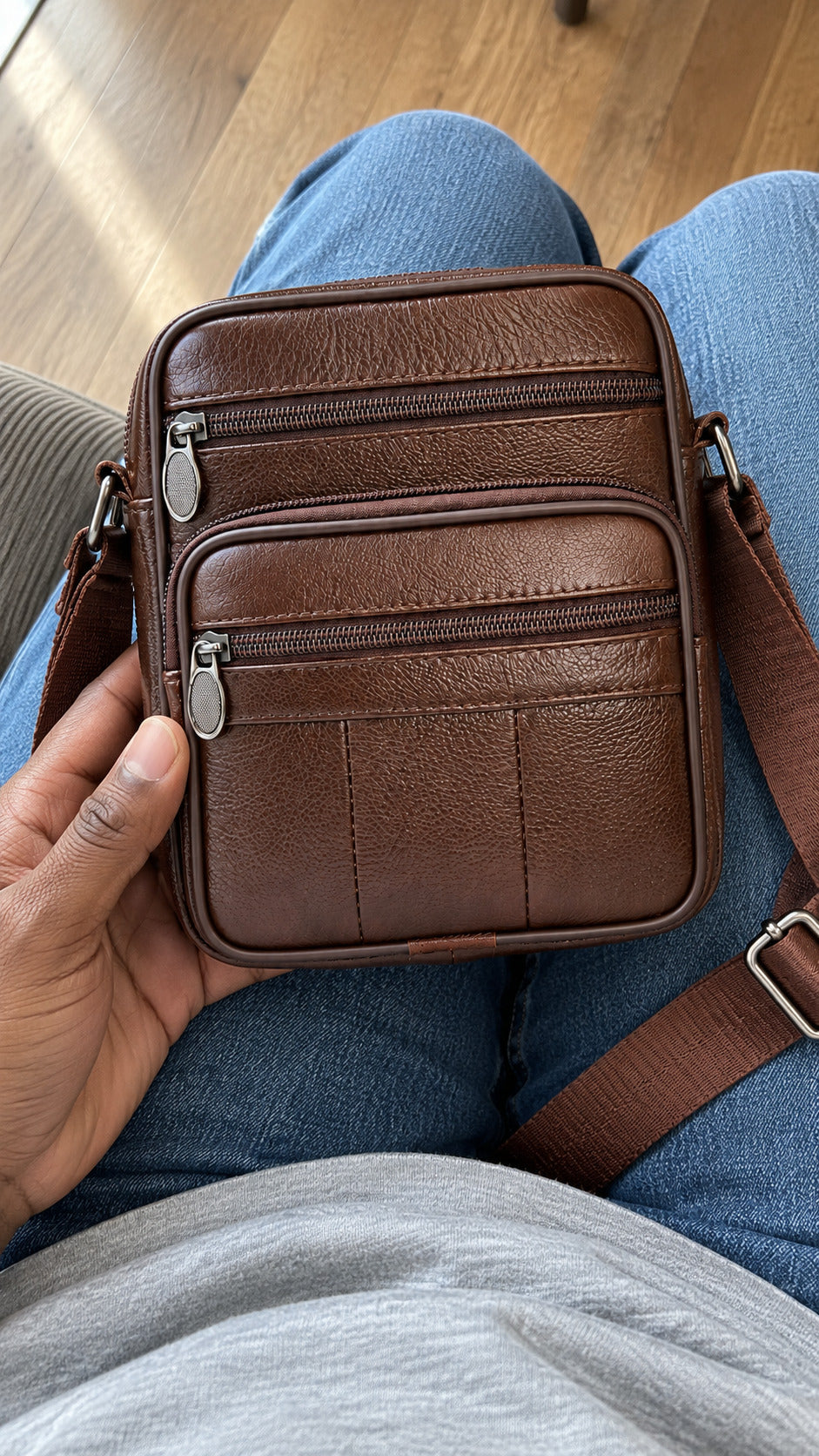 A brown leather crossbody phone bag with multiple zippered compartments and a shoulder strap, held by a person wearing blue jeans and a gray sweatshirt.