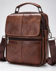 A brown textured leather crossbody bag with a top handle, front zippered pocket, and adjustable shoulder strap, photographed on a light gray studio background.