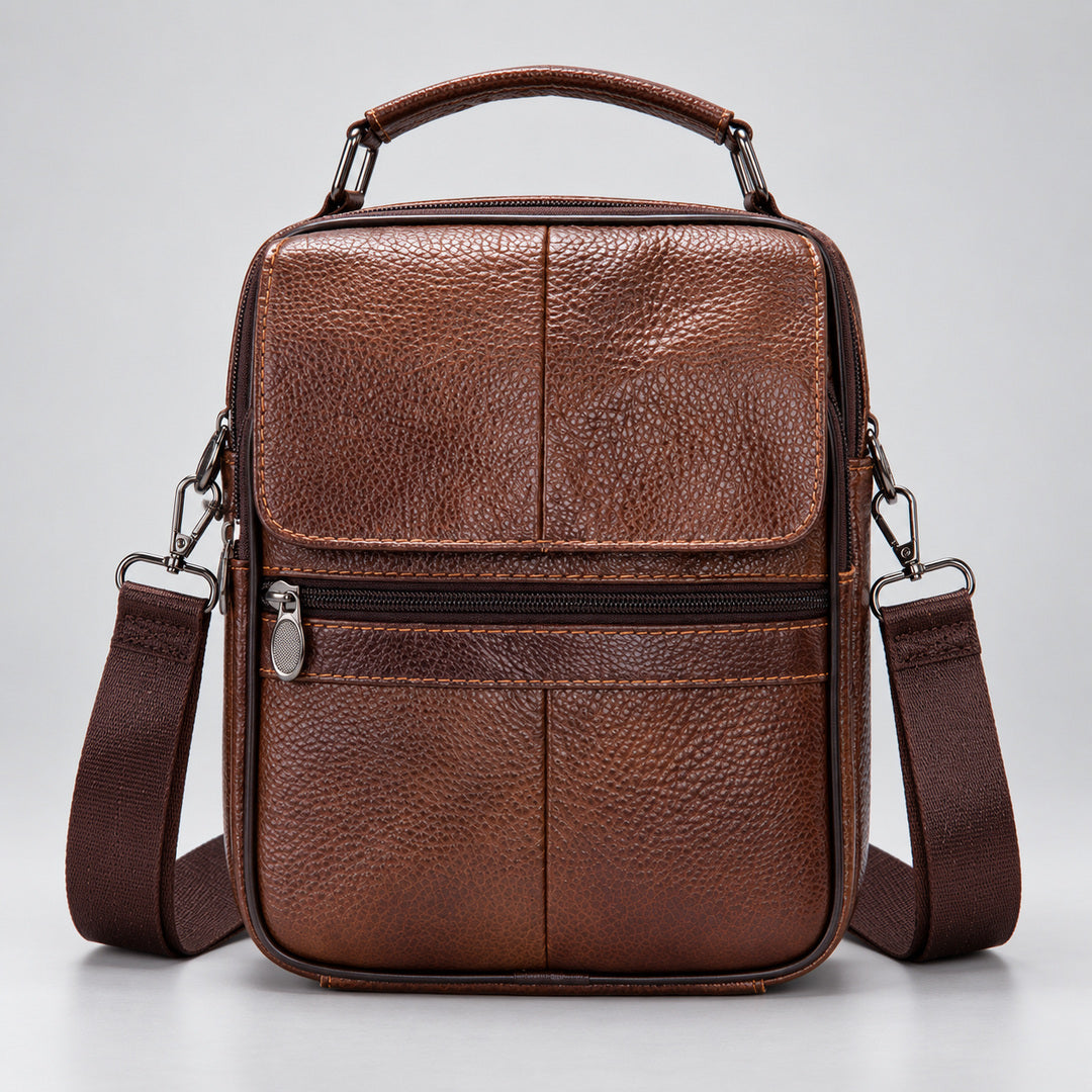 A brown textured leather crossbody bag with a top handle, front zip pocket, and matching shoulder strap, photographed against a light gray studio background.