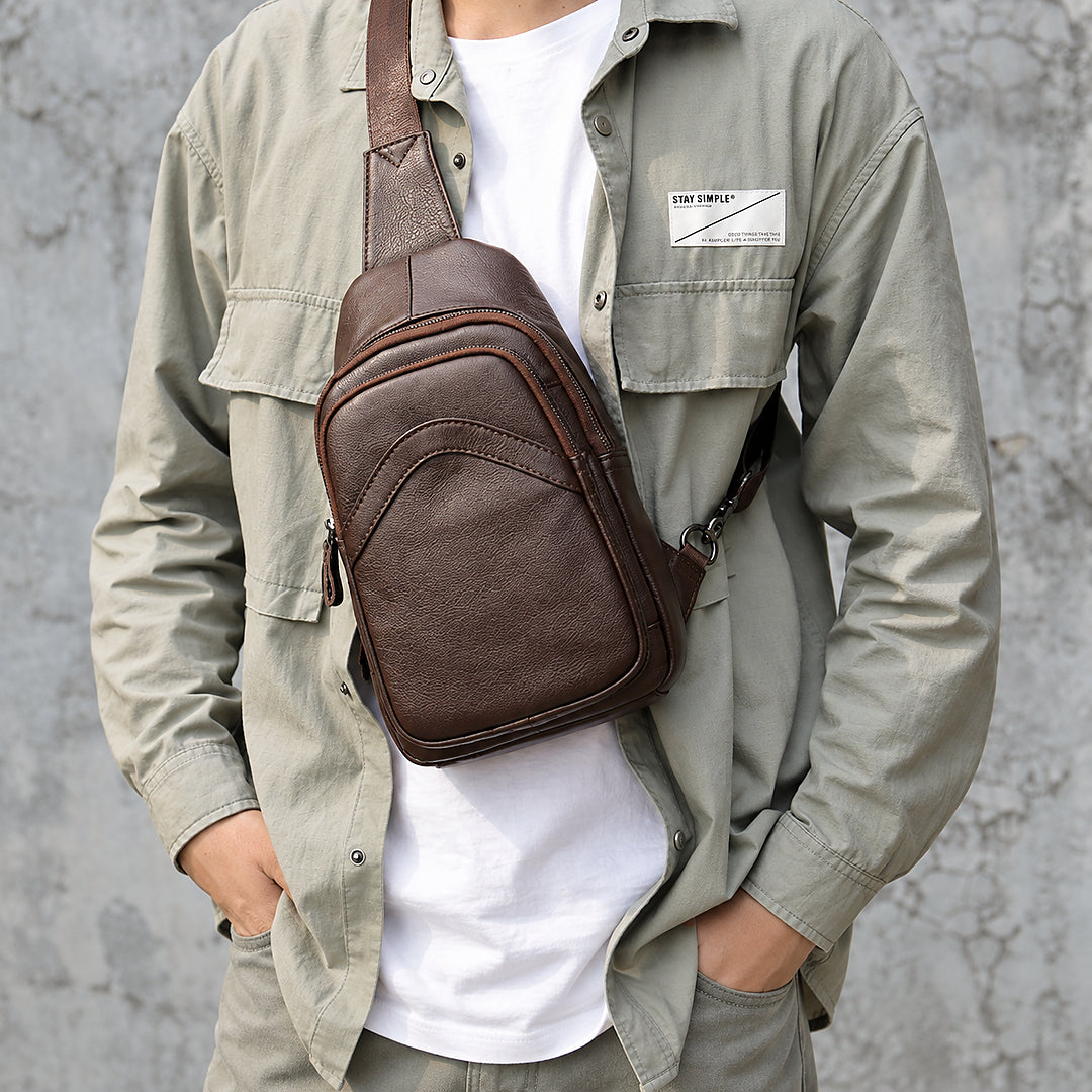 A man wearing a brown leather crossbody sling bag across his chest, styled with an olive green overshirt and white t-shirt against a textured gray wall.