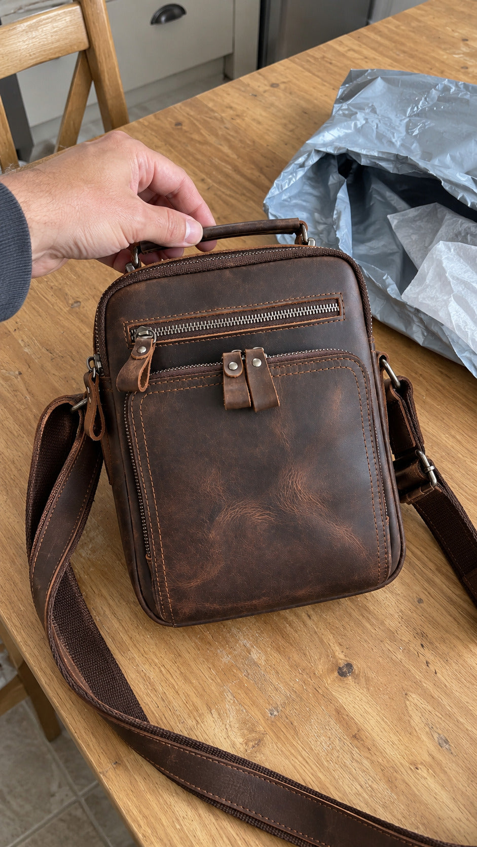 A brown distressed leather crossbody bag with a top handle, front zip pocket, and adjustable shoulder strap, shown on a wooden table with a hand holding the handle and a gray shipping bag in the background.