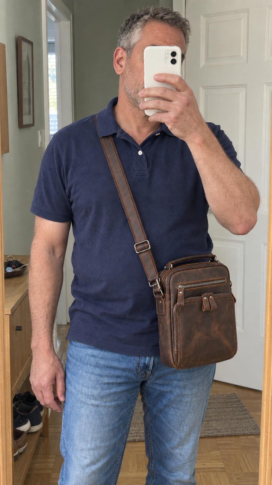 A man takes a mirror selfie wearing a brown leather crossbody bag with a wide adjustable strap, styled with a navy blue polo shirt and blue jeans.