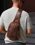 Men wearing a brown leather sling bag over his shoulder on a city street.