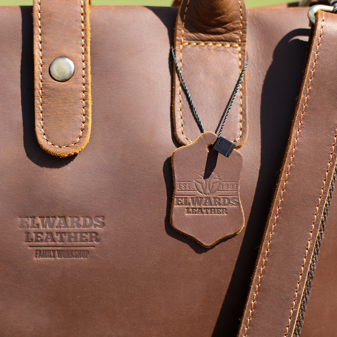 Men&#39;s leather work bag with Elwards Leather logo and detachable strap resting on a wooden surface in natural light.