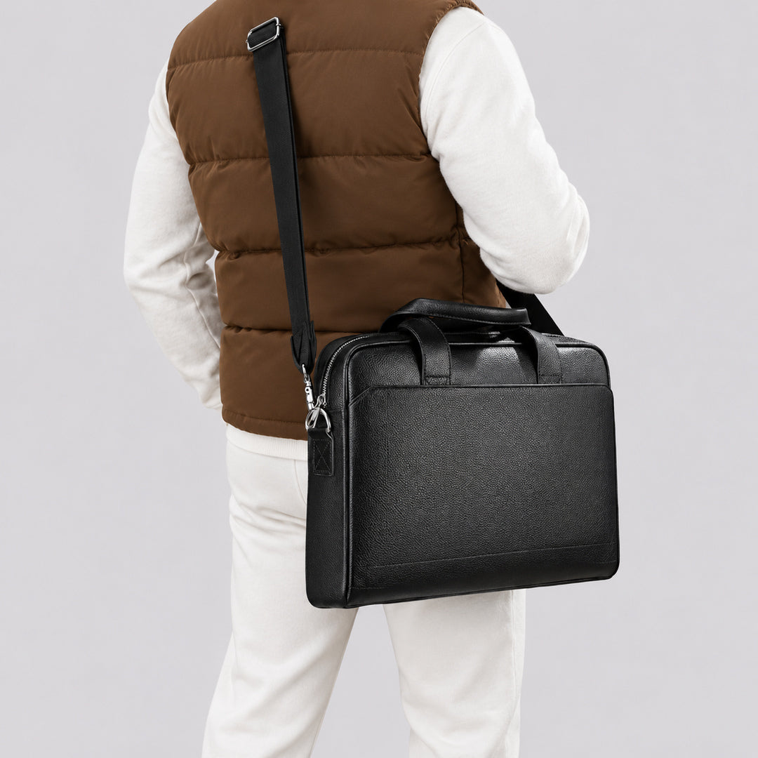 A man wearing a brown puffer vest, white long-sleeve shirt, and white pants is shown from the back while carrying a black leather briefcase-style shoulder bag with a top handle and adjustable strap against a light gray studio background.