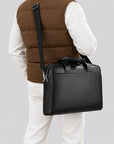 A man wearing a brown puffer vest, white long-sleeve shirt, and white pants is shown from the back while carrying a black leather briefcase-style shoulder bag with a top handle and adjustable strap against a light gray studio background.