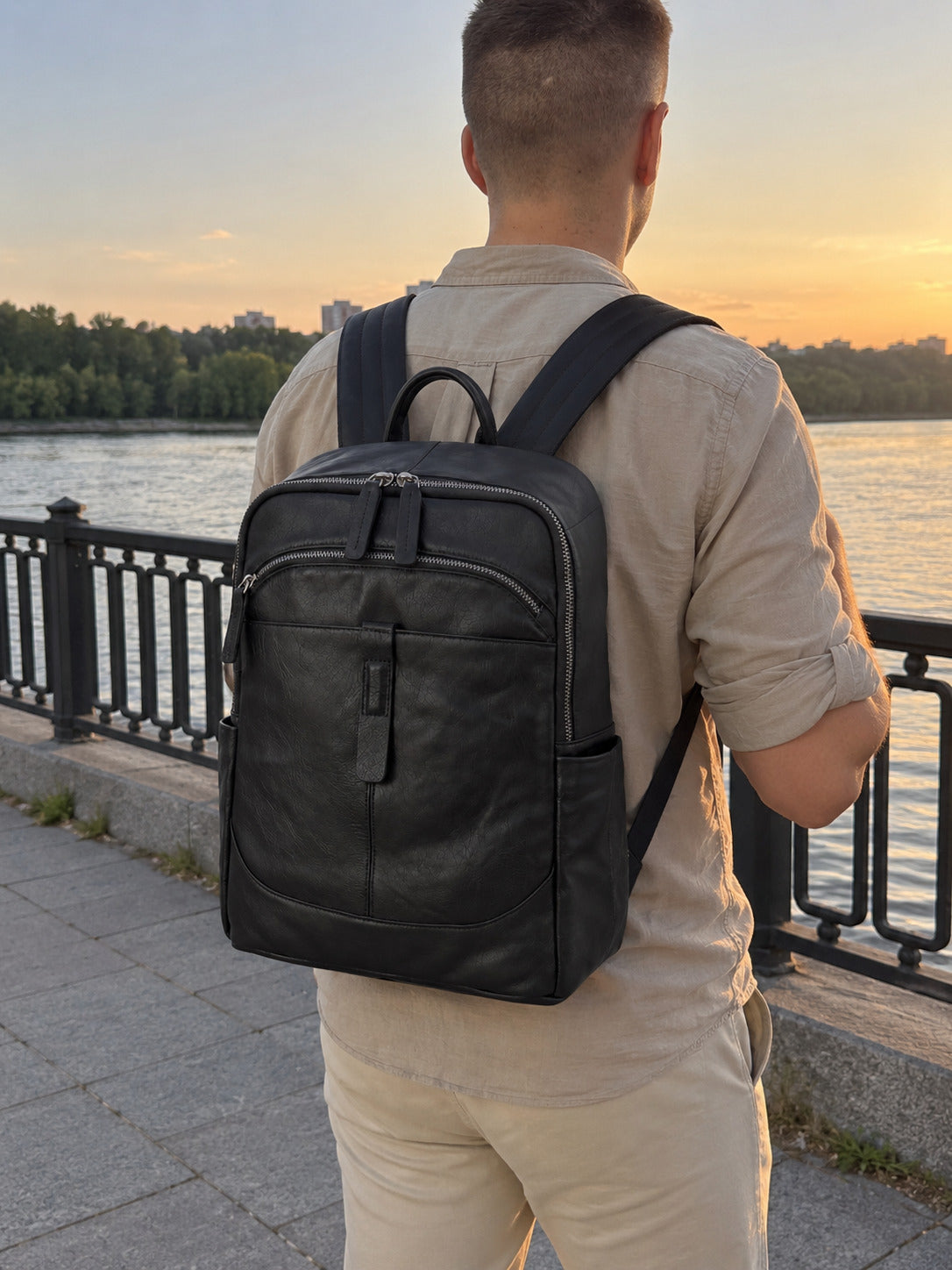 A man viewed from behind wearing a black leather backpack while standing on a riverside walkway at sunset.