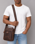 A man wearing a white t-shirt and blue jeans carries a brown leather crossbody bag with a top handle and adjustable shoulder strap against a solid gray background.