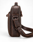 A dark brown distressed leather crossbody bag with multiple zippered compartments and an adjustable shoulder strap, shown from a side profile view on a white background.