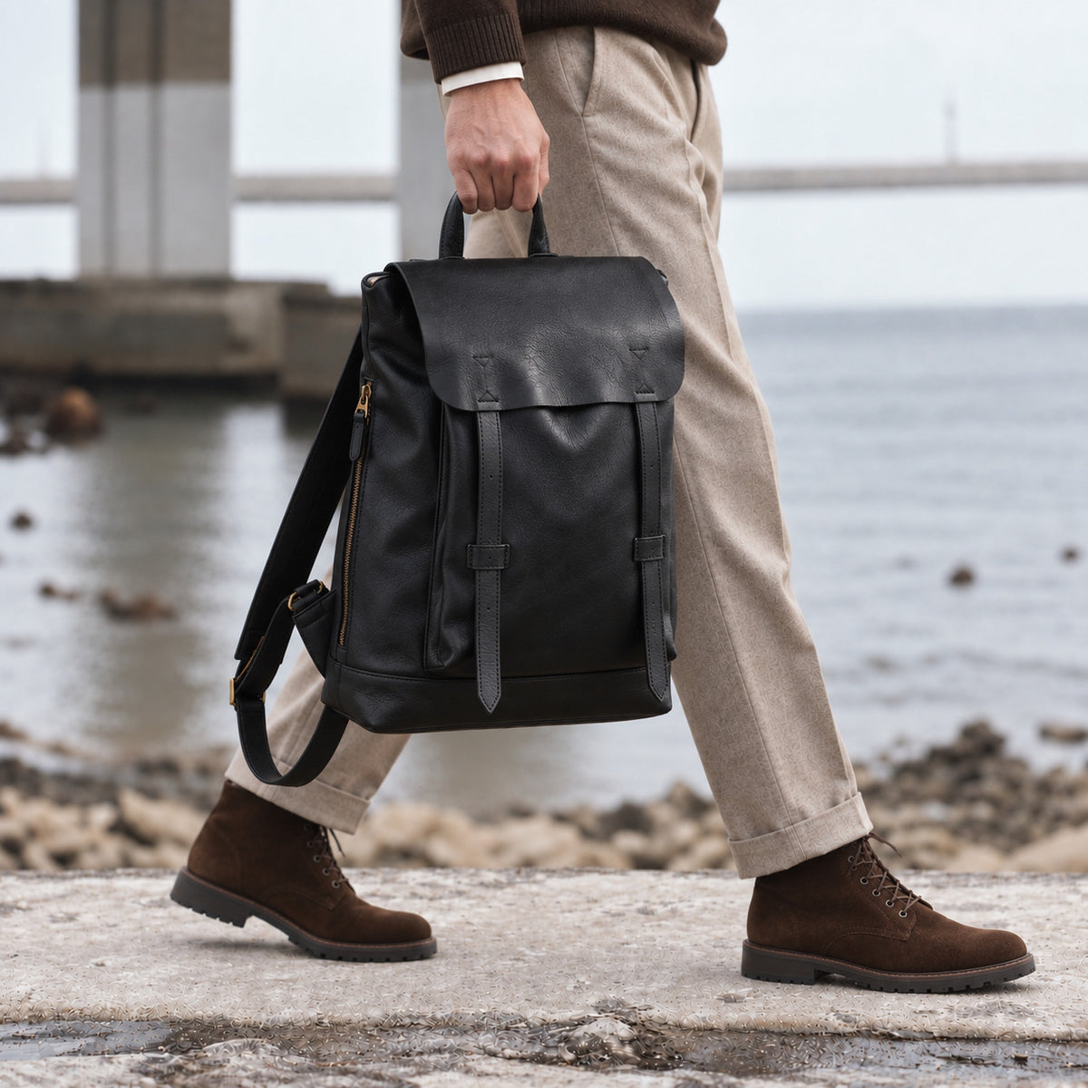 Minimalist black leather backpack for men with flap closure, top carry handle and adjustable shoulder straps, carried by hand