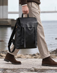 Minimalist black leather backpack for men with flap closure, top carry handle and adjustable shoulder straps, carried by hand