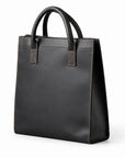 Minimalist black full-grain leather tote at an angle showing its tall shape and stitched side panel