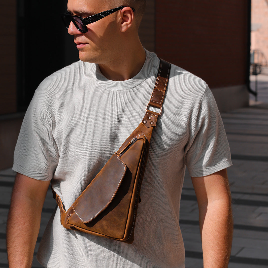 Man wearing a brown leather crossbody bag and sunglasses outdoors.