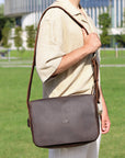 Model wearing a men's brown leather satchel bag in an outdoor urban setting