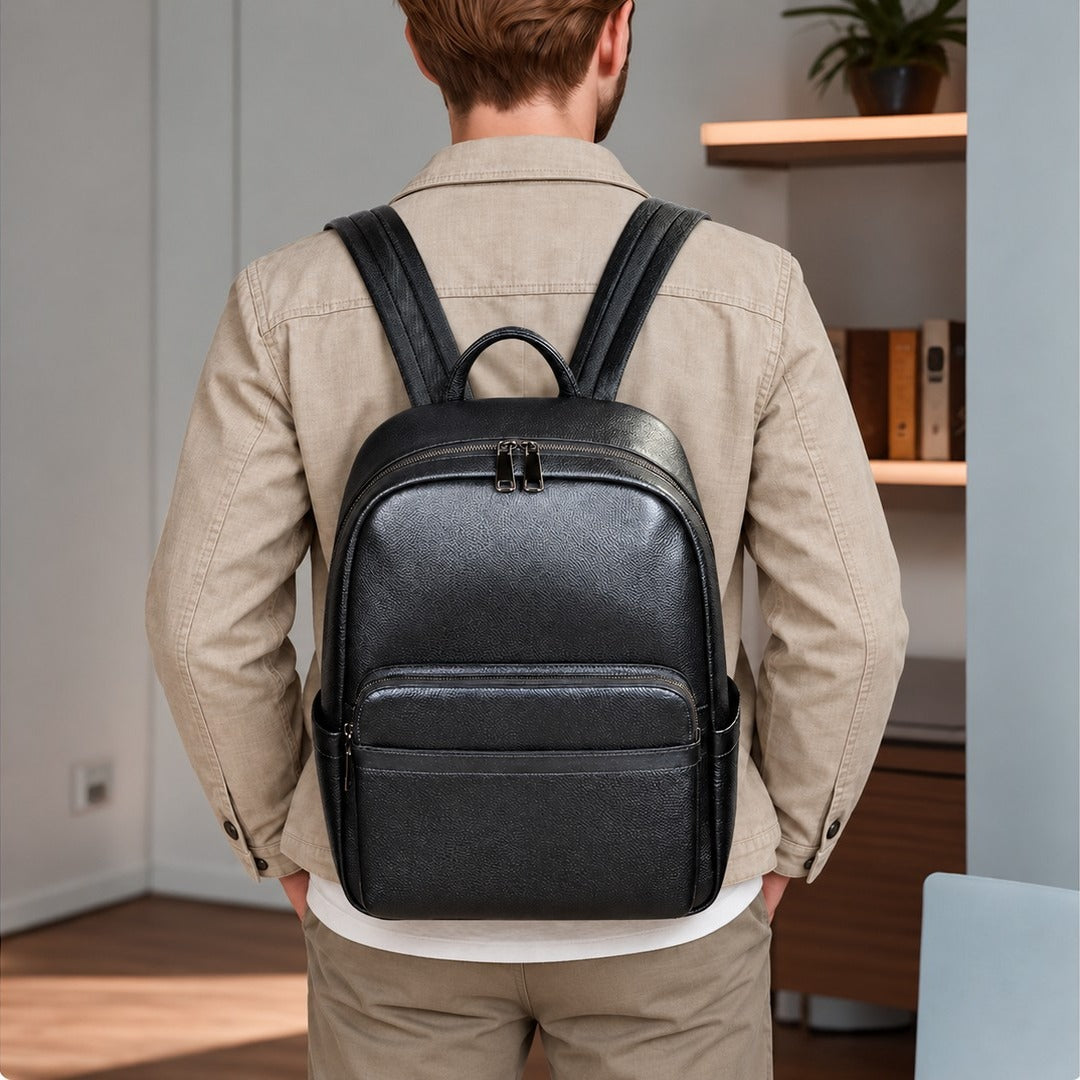 A man viewed from behind wearing a black textured leather backpack on his shoulders, dressed in a beige jacket and trousers in an indoor office setting.