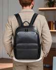A man viewed from behind wearing a black textured leather backpack on his shoulders, dressed in a beige jacket and trousers in an indoor office setting.