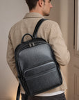 A man wearing a beige jacket is shown from the back, carrying a black textured leather backpack with top handles and dual zipper compartments.