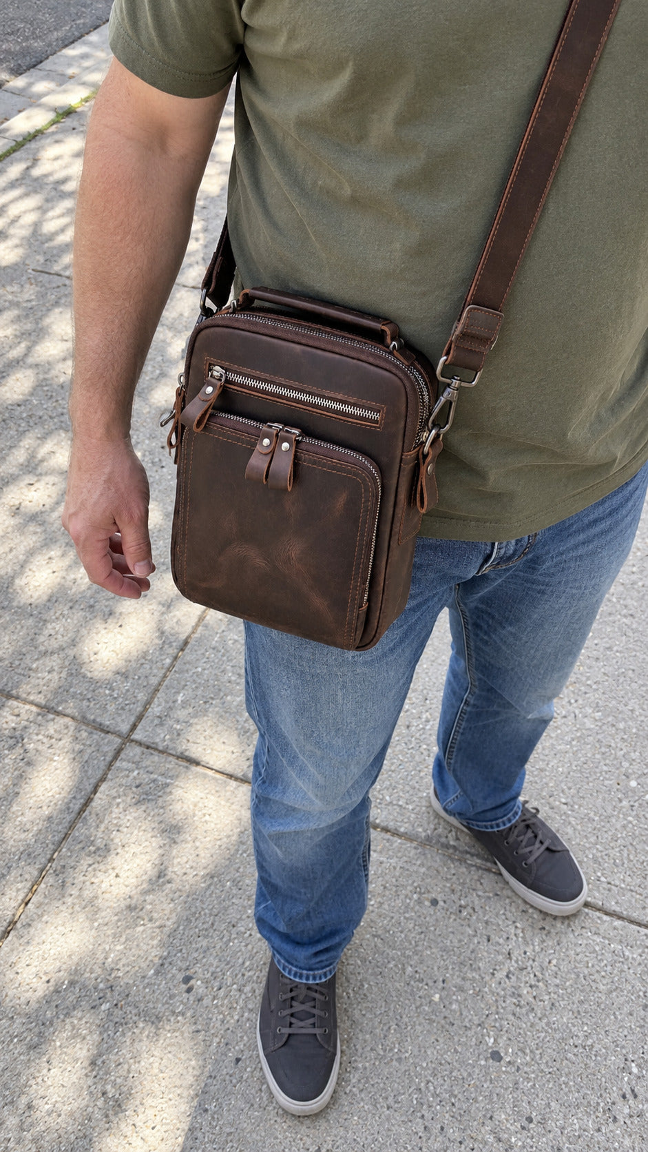 A man wearing a green t-shirt, blue jeans, and gray sneakers carries a brown leather crossbody bag on his shoulder while standing on a concrete sidewalk.