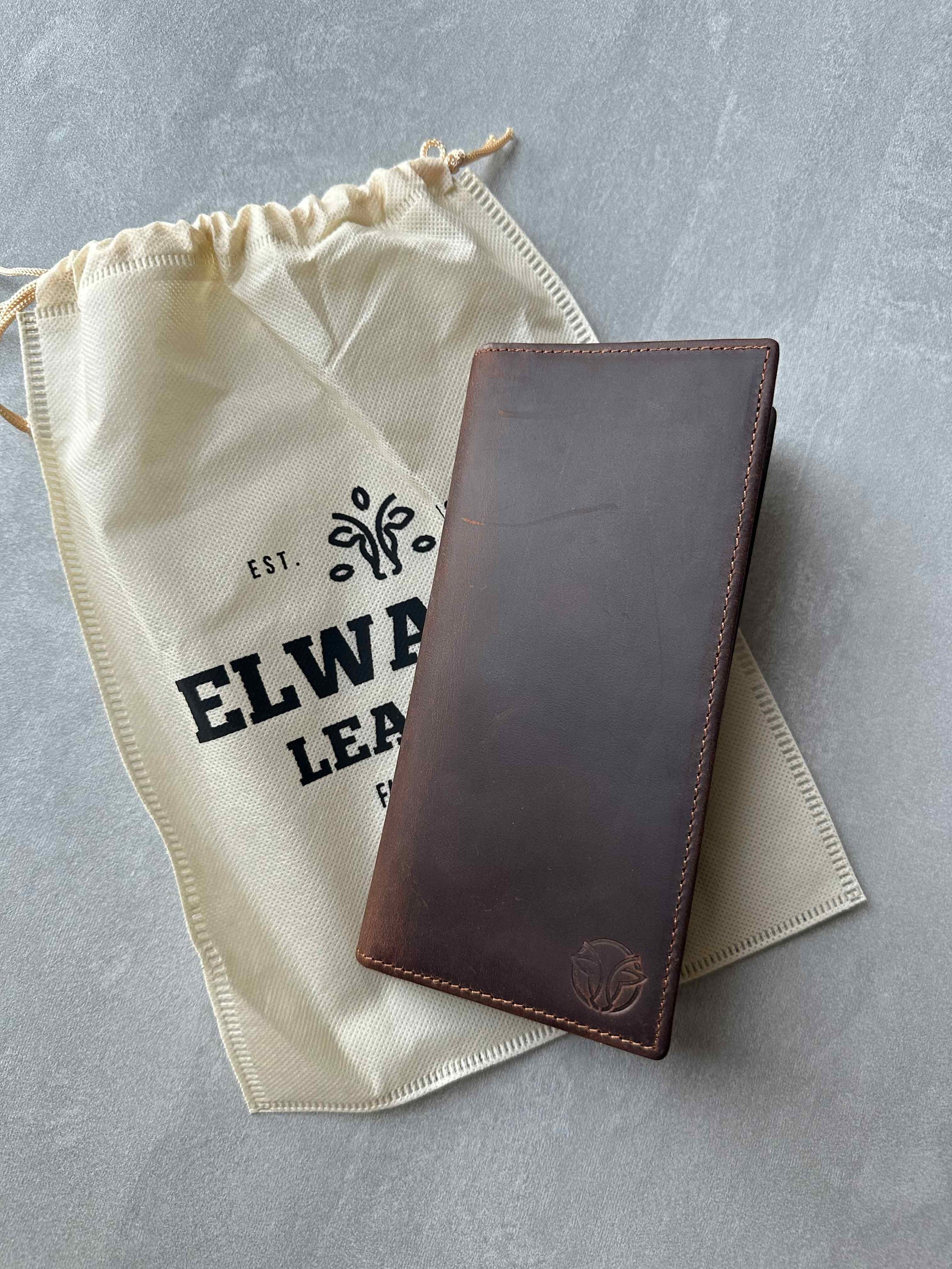 Brown leather wallet on a gray surface with an Elwards Leather drawstring bag.