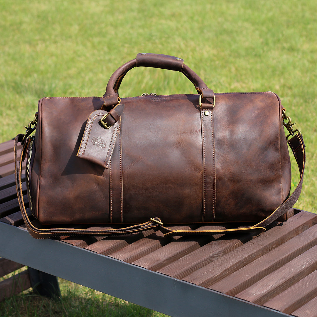 Premium brown leather travel bag displayed on a wooden bench with a detachable shoulder strap.