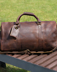 Premium brown leather travel bag displayed on a wooden bench with a detachable shoulder strap.