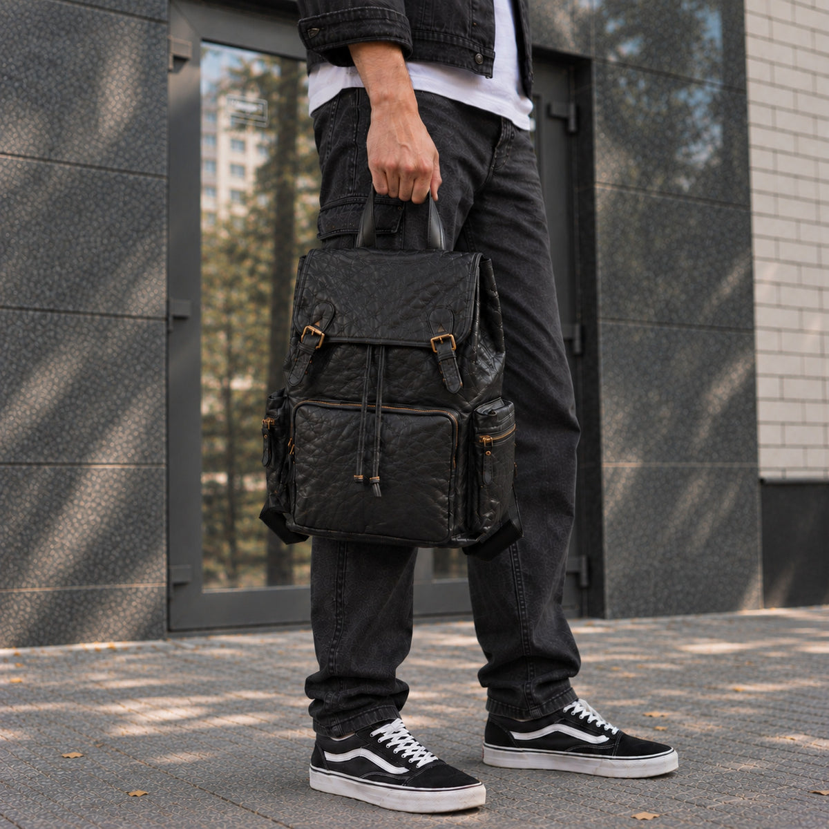 Quilted black leather travel backpack for men with buckle flap, front zip pocket, side pockets and top carry handle