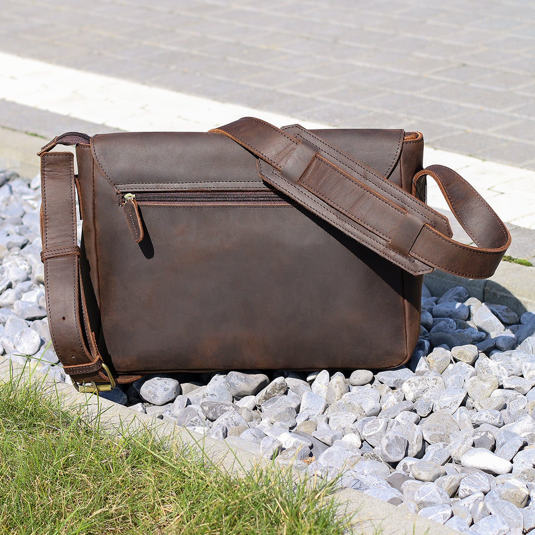 Rear view of a leather satchel on gravel with a zippered pocket and adjustable strap