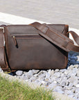 Rear view of a leather satchel on gravel with a zippered pocket and adjustable strap