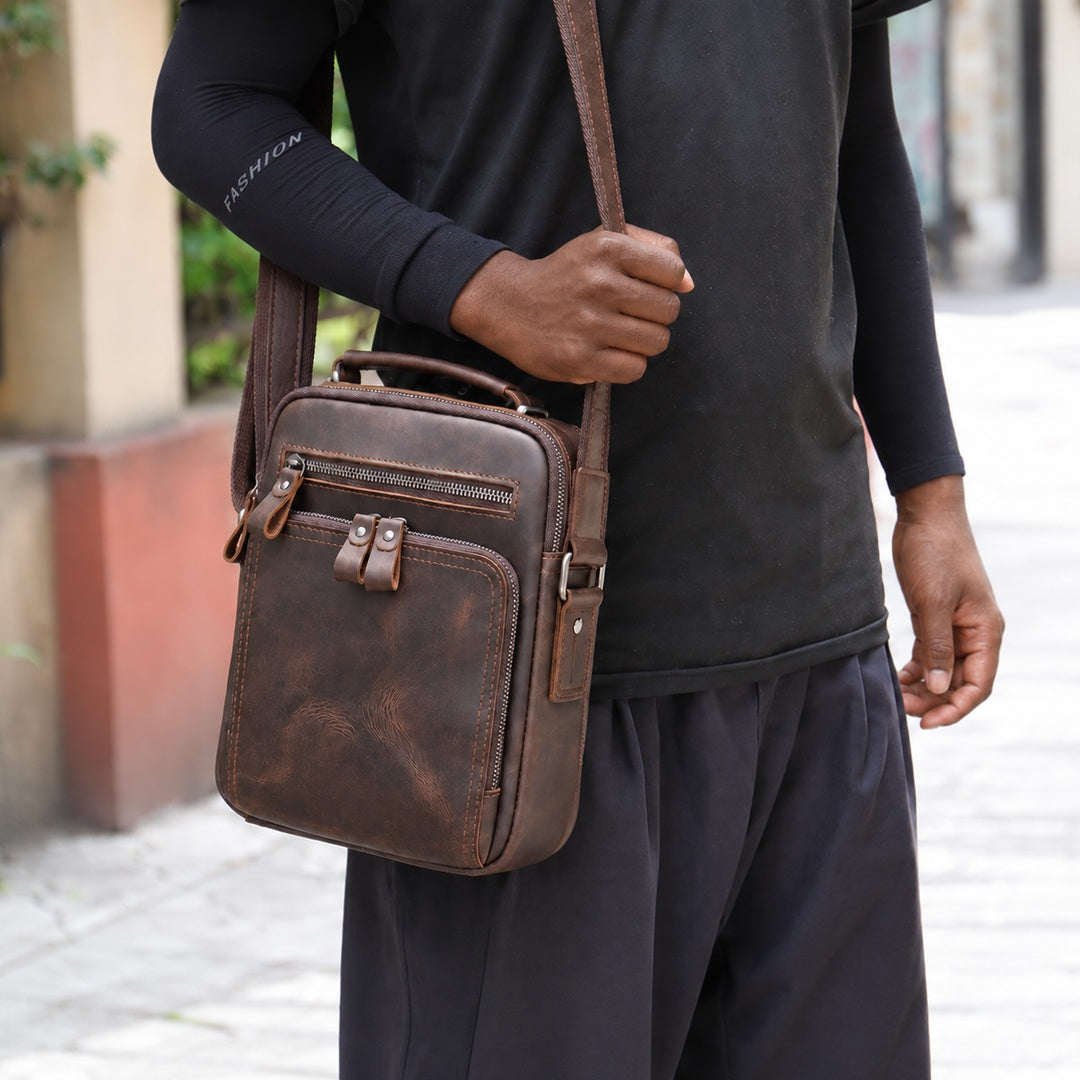 A man wearing a black long-sleeve shirt and dark pants carries a brown distressed leather crossbody bag over his shoulder in an outdoor setting.