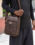 A man wearing a black long-sleeve shirt and dark pants carries a brown distressed leather crossbody bag over his shoulder in an outdoor setting.