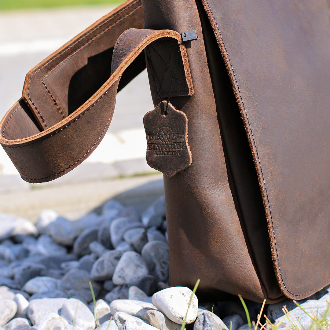 Side profile of a dark brown men&#39;s leather shoulder bag resting on gravel, showcasing tag and clean stitching