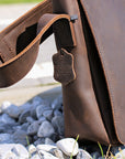 Side profile of a dark brown men's leather shoulder bag resting on gravel, showcasing tag and clean stitching