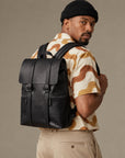 Sleek black leather backpack for men with flap top, vertical strap details and front zip pockets, worn as an urban everyday bag