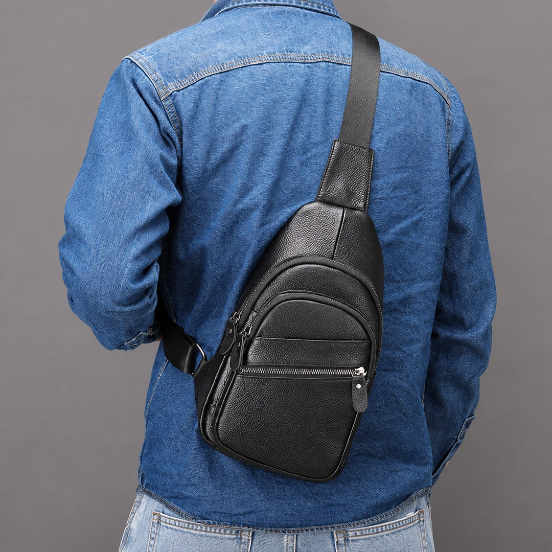 A man wearing a blue denim shirt and jeans, shown from the back, wearing a black textured leather sling bag across his back. The bag has a single adjustable strap and a front zippered pocket.