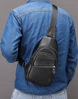 A man wearing a blue denim shirt and jeans, shown from the back, wearing a black textured leather sling bag across his back. The bag has a single adjustable strap and a front zippered pocket.