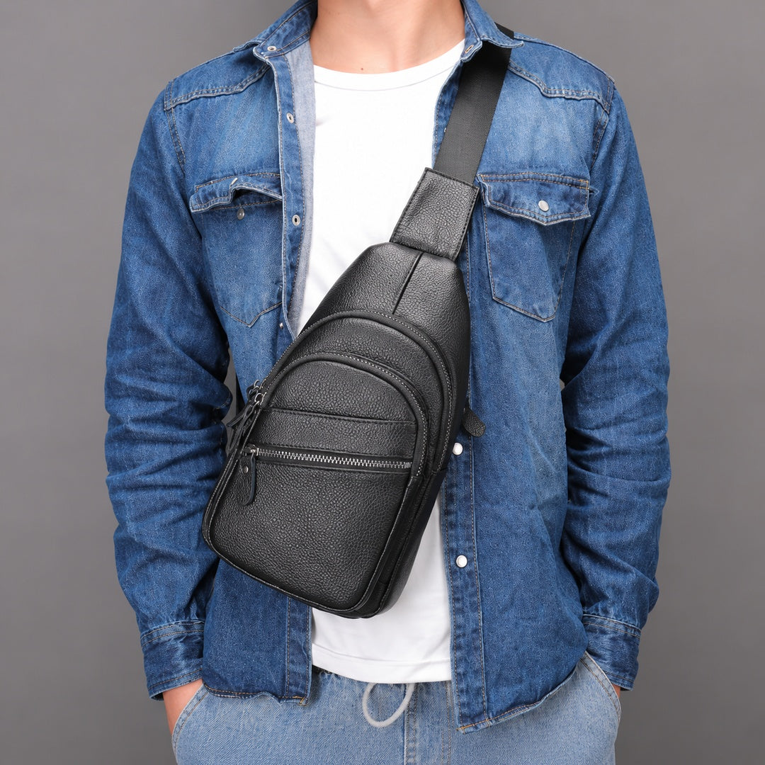 A black textured leather sling bag with a wide adjustable shoulder strap, worn crossbody over a blue denim shirt and white t-shirt, against a solid gray studio background.