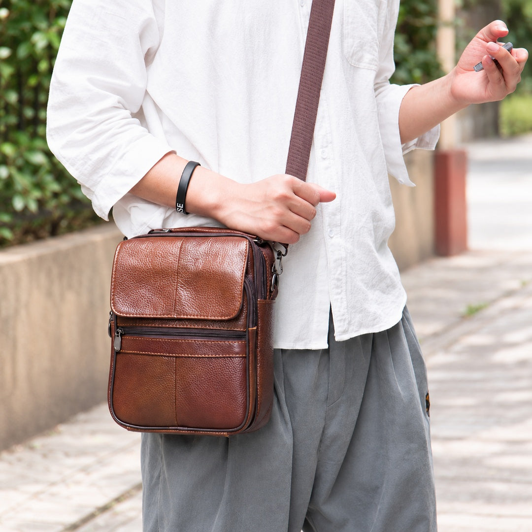 A man wearing a white button-up shirt and gray pants carries a brown leather crossbody bag with a matching strap in an outdoor setting.