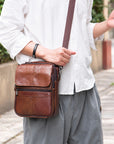 A man wearing a white button-up shirt and gray pants carries a brown leather crossbody bag with a matching strap in an outdoor setting.