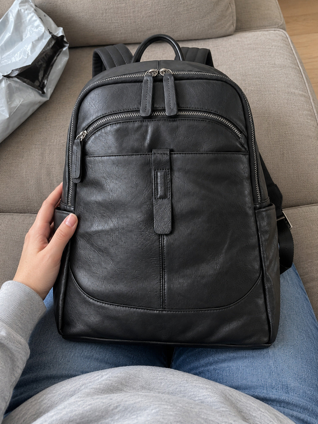 A black leather backpack resting on a person's lap on a beige sofa, with a hand holding the side of the bag.