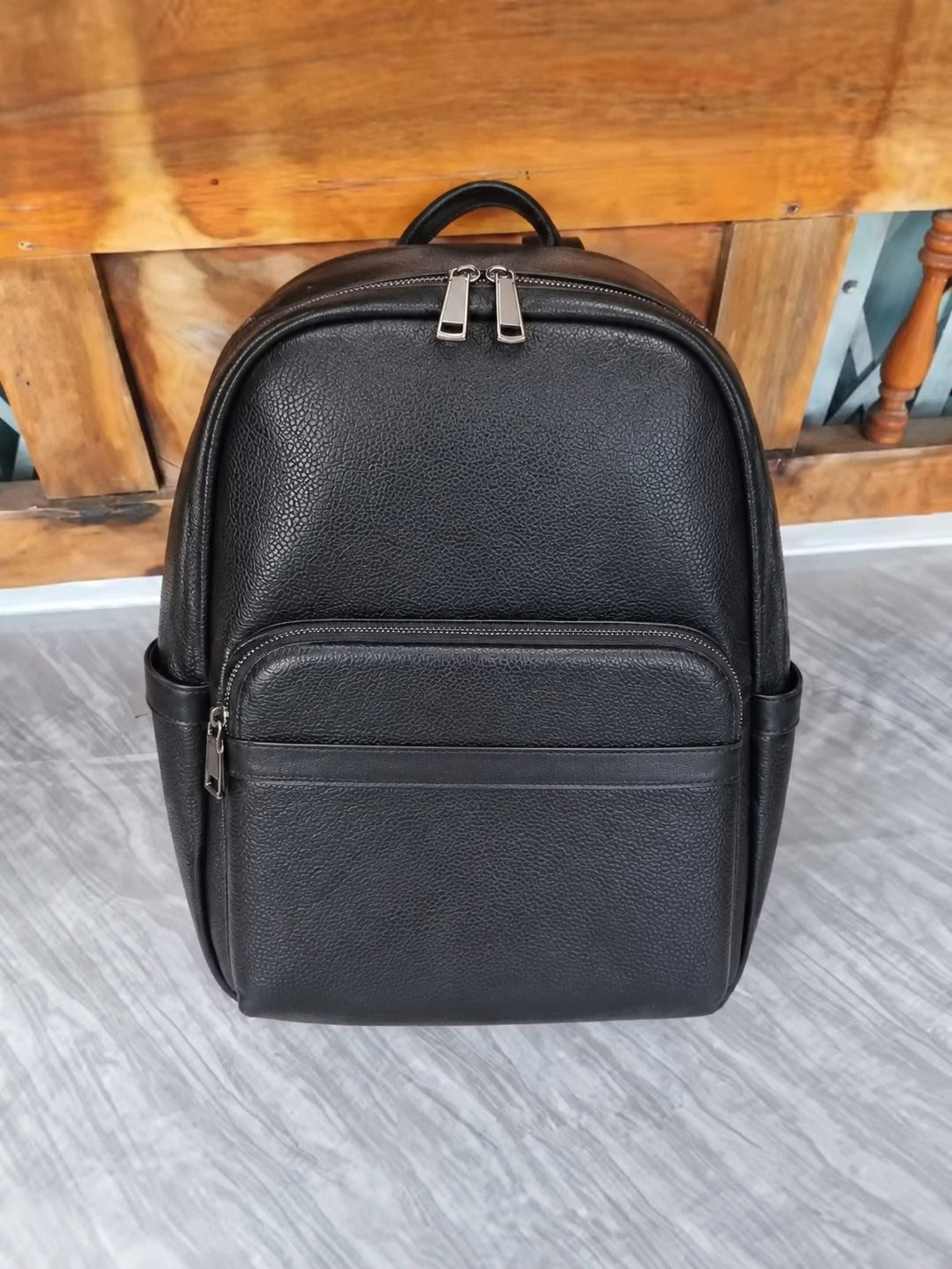 A black pebbled leather backpack with dual zipper pulls, shown standing on a gray floor in front of a wooden bench.
