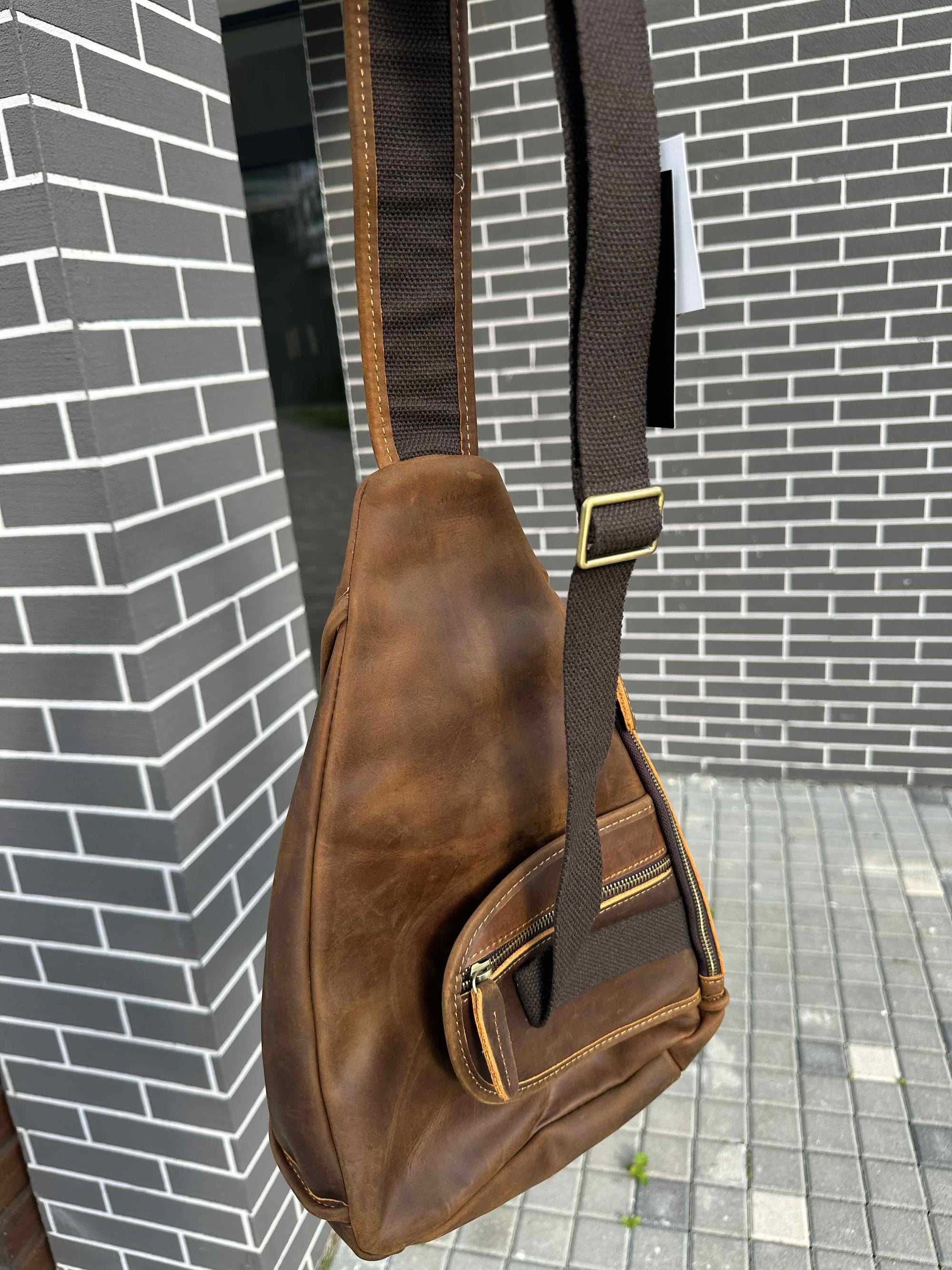 Brown leather sling bag with a strap against a brick wall.