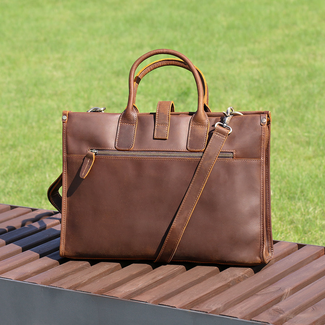 Back view of slim brown men&#39;s vintage leather work bag with detachable strap and back zipper pocket on a wooden bench.