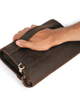 Hand gripping the back strap of a men's dark brown leather wrist bag, highlighting its comfort and secure design.